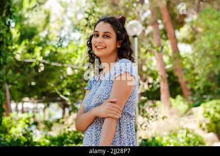 Ritratto di donna bruna carina indossando un abito estivo floreale blu in piedi sul parco della città, all'aperto guardando la macchina fotografica mentre si posa. Bellezza naturale conc Foto Stock