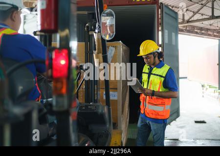 Conducente carrello elevatore asiatico che carica un contenitore di carico di spedizione con un pallet completo con le scatole nel terminal del porto logistico. Il lavoratore di magazzino asiatico e la sicurezza i Foto Stock