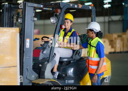 Uomo asiatico insegnamento donna conducente guida carrello elevatore auto in industria con sorriso, capacità di ragazza e diversità di carriera indossare casco in giubbotto riflettente Foto Stock