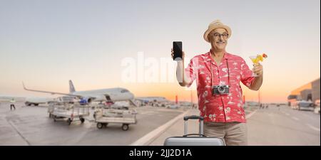 Turista maschio maturo con una valigia che tiene un cocktail e uno smartphone su un grembiule aeroporto Foto Stock