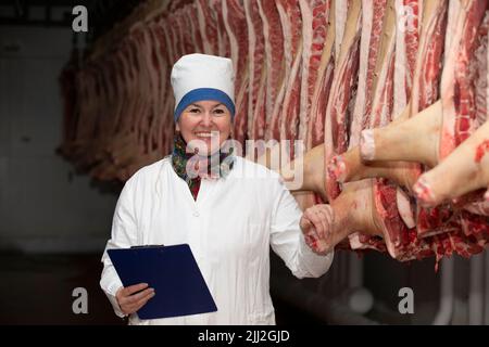 Operatore dello stabilimento di imballaggio delle carni di fronte alle carcasse macellate. Produzione di carne. Un macellaio professionista tra file di carcasse di suino, guardando il venuto Foto Stock