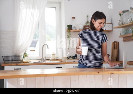 Una donna in cucina al computer con un sorriso. Una donna digita su un computer, lavora da casa o comunica sui social network. Una donna beve il tè e tiene una tazza in mano. Foto di alta qualità Foto Stock