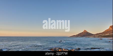 Vista sull'oceano dell'acqua di mare su una spiaggia rocciosa, montagne e un cielo blu con spazio copia di Lions Head a Città del Capo, Sud Africa. Calma marea, serena e. Foto Stock