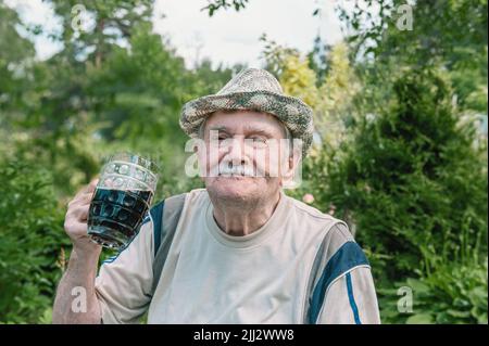 grande ritratto di un vecchio uomo dai capelli grigi in un cappello con un bicchiere di bevanda scura. l'uomo con una tazza di vetro è seduto in giardino. Spazio di copia. Foto Stock