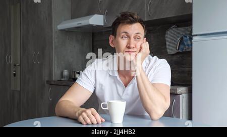 L'uomo assonnato si assapiglia al tavolo e beve una tazza di caffè in cucina Foto Stock