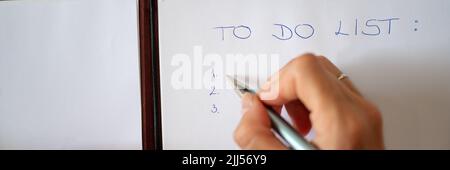 Immagine grandangolare della mano femminile che scrive una lista di cose da fare su un foglio bianco di carta. Vista dall'alto. Foto Stock