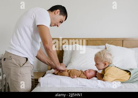 Ragazzo che guarda il padre che abbottonava le rompper del bambino sul letto Foto Stock