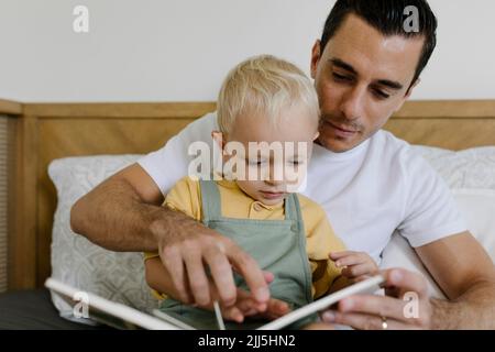Uomo che legge libro con figlio seduto sul letto a casa Foto Stock