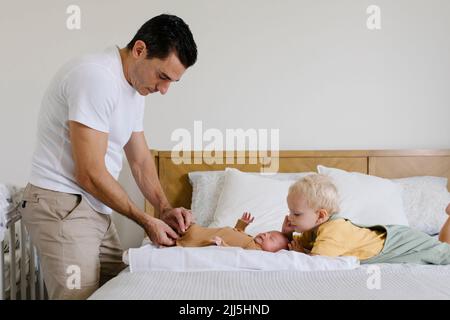 Ragazzo che guarda l'uomo che abbottonano le rompper del bambino sul letto a casa Foto Stock