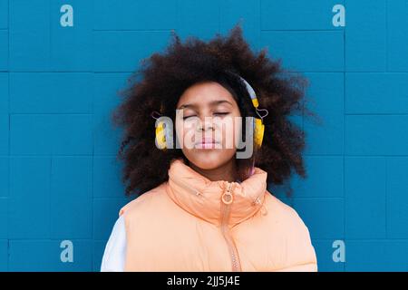 Ragazza con gli occhi ha chiuso l'ascolto della musica attraverso le cuffie wireless di fronte alla parete blu Foto Stock