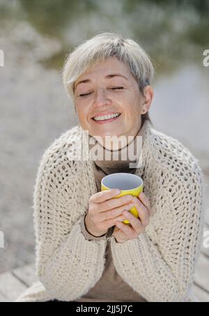 Donna matura sorridente con occhi chiusi che tiene la tazza Foto Stock