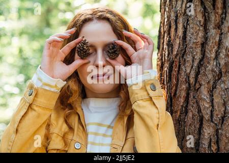 Donna sorridente che copre gli occhi con cono di pino nella foresta Foto Stock