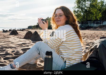 Giovane donna che tiene una tazza seduta in spiaggia Foto Stock