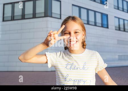 Ragazza felice che mostra segno di pace in piedi di fronte alla costruzione Foto Stock