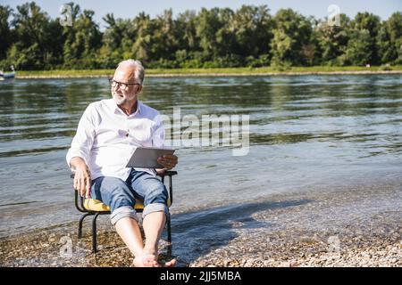 Uomo anziano che tiene un tablet PC seduto su una sedia sul lungofiume Foto Stock