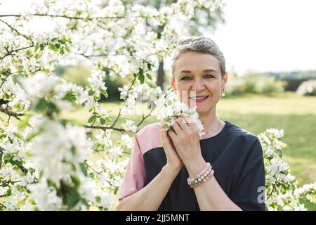 Donna sorridente con fiore bianco in piedi in giardino Foto Stock