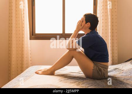 Donna despairata che ricopre il viso con le mani sedute sul letto di casa Foto Stock