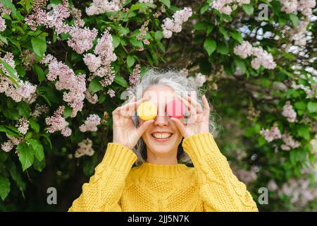 Donna felice che copre il viso con macaroon di fronte al lilla al parco Foto Stock
