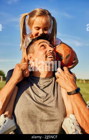 Uomo felice guardando la figlia seduta con palla di rugby sulle spalle Foto Stock