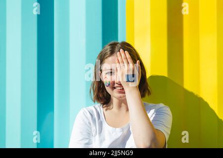 Ragazza sorridente adolescente con vernice simbolo di pace sul viso che copre l'occhio di fronte al blu e giallo parete Foto Stock