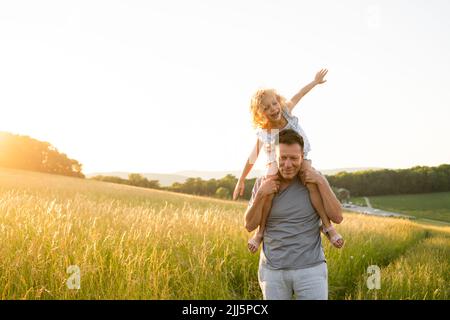 Uomo sorridente che porta la figlia seduta con le braccia tese al campo Foto Stock