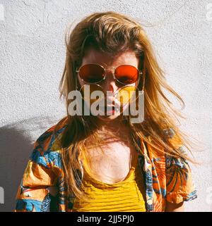 Donna con capelli marroni che indossano occhiali da sole rossi e gialli davanti alla parete Foto Stock