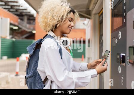 Donna sorridente che scansiona il codice QR tramite smartphone sul distributore automatico di biglietti Foto Stock