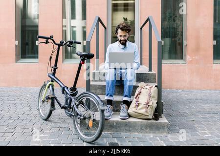 Uomo d'affari che utilizza un computer portatile seduto su gradini in bicicletta Foto Stock