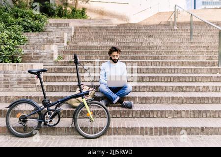 Freelance che utilizza un computer portatile seduto su gradini vicino alla bicicletta Foto Stock