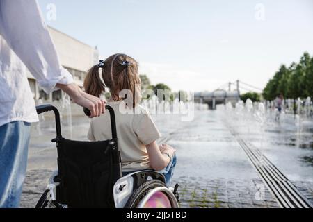 Madre in piedi dietro la figlia con disabilità seduta in sedia a rotelle al parco il giorno di sole Foto Stock