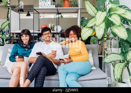 Sorridenti colleghi multirazziali con smartphone sul divano in ufficio Foto Stock