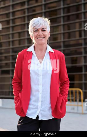 Donna d'affari sorridente con giacca rossa davanti all'edificio Foto Stock