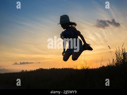 Silhouette di una ragazza in un cappello, una piccola strega vola al crepuscolo di Halloween sullo sfondo del cielo del tramonto. Salto tra cielo e terra per la gioia Foto Stock