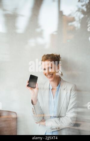 Donna d'affari felice che mostra il telefono cellulare in un caffè visto attraverso il vetro Foto Stock