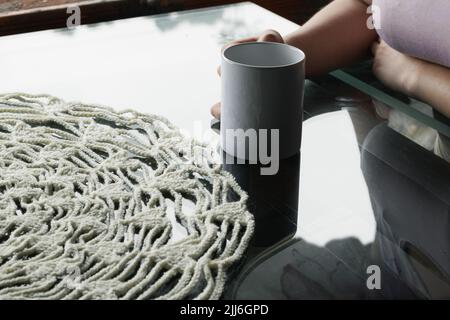 mano della donna latina che tiene una tazza bianca su un tavolo di vetro, con un centrotavola bianco rotondo. ragazza rilassata nel suo ufficio bevendo una tazza di caffè Foto Stock