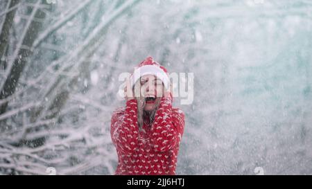La donna allegra gode wonderland invernale. La neve vola su di lei ed è felice, gioisce il freddo. Ragazza in rosso maglione di Natale e cappello Santa. Foto Stock