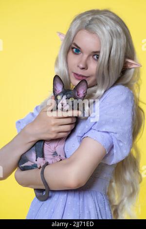 Giovane donna cosplay elf in abito estivo blu, con capelli lunghi biondi e occhi di diversi colori che tengono in mano Sphinx gattino, guardando la macchina fotografica Foto Stock