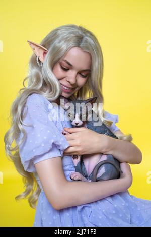 Giovane donna cosplay elf in abito blu sorride felicemente, abbracciando delicatamente Sphinx gattino al petto e guardandolo giù. Elf ha capelli biondi meravigliosi Foto Stock
