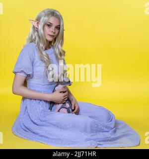 Giovane donna bionda cosplay elf con occhi di colori diversi si siede su sfondo giallo, tiene Sfinx gatto sul giro, guarda la macchina fotografica Foto Stock