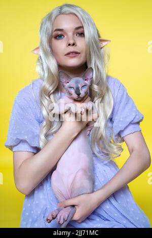 Coscrayer Elf giovane donna che tiene Sfinge gatto nelle sue mani e che lo mostra su sfondo giallo. Elf in abito blu, con capelli biondi ricci lunghi Foto Stock