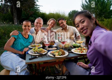 Gruppo di amici che si divertono durante la festa estiva. Donna asiatica che prende selfie all'ora della cena del barbecue. Persone di mezza età che gelano fuori mangiare e. Foto Stock