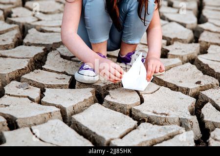 La ragazza abbassa la barca di carta sul terreno asciutto e incrinato. Crisi idrica e concetto di cambiamento climatico. Riscaldamento globale Foto Stock