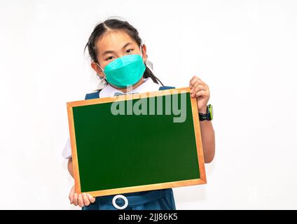 Ritratto asiatico studente bambino in uniforme identificata scuola, tenendo piccola lavagna o lavagna, indossare maschera viso, immagine isolata su bianco backgrou Foto Stock