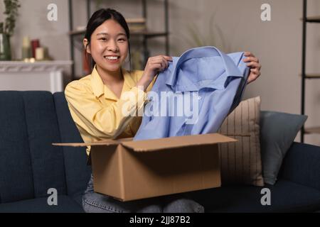 Happy Korean Woman Holding New Shirt Unpacking Box sitting Indoor Foto Stock