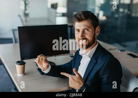 Uomo d'affari felice in vestito che tiene il bitcoin d'oro e punta il dito su di esso, seduto sul posto di lavoro con il computer Foto Stock