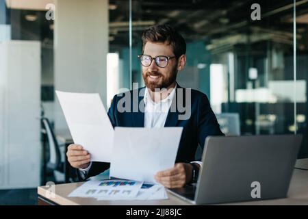 Felice uomo d'affari che legge i documenti e le relazioni di affari, rallegrandosi negli affari dell'azienda, seduto in ufficio moderno Foto Stock
