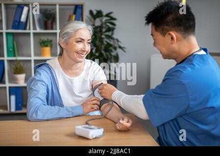 Felice giovane uomo medico asiatico in uniforme misura la pressione sanguigna con tonometro a pazienti anziani europei Foto Stock