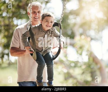 Ragazzo caucasico eccitato che si diverte mentre si siede nell'altalena ed è spinto dal nonno al parco. Uomo anziano attivo che lega e gioca Foto Stock