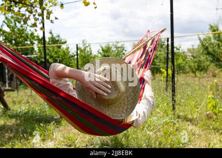 vista posteriore della donna in giorno d'estate che indossa cappello prendendo un pisolino nell'amaca nel suo cortile rilassante durante le vacanze Foto Stock