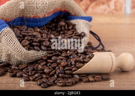 Chicchi di caffè nero arrostiti con un cucchiaio di legno, fuoriuscite dal sacco sul tavolo. Foto per una caffetteria, un caffè o un ristorante. Foto Stock